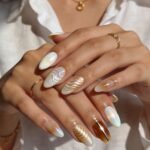 Hands with almond-shaped nails featuring elegant 3d nail art, including pearlescent chrome finishes and wavy textures in white and gold, with accents of amber tones and metallic embellishments.
