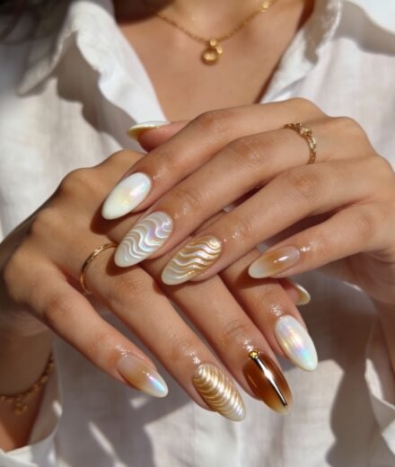 Hands with almond-shaped nails featuring elegant 3d nail art, including pearlescent chrome finishes and wavy textures in white and gold, with accents of amber tones and metallic embellishments.