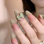Close-up of a hand with almond-shaped nails featuring a sheer pink base, light green lace-like tips, and delicate embellishments including a satin green bow and small gems for an elegant and vintage-inspired look.
