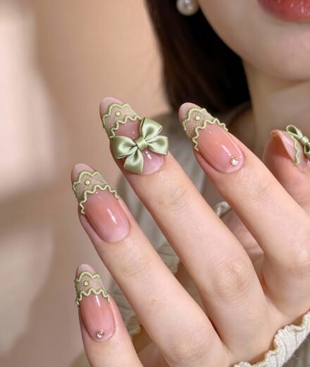 Close-up of a hand with almond-shaped nails featuring a sheer pink base, light green lace-like tips, and delicate embellishments including a satin green bow and small gems for an elegant and vintage-inspired look.