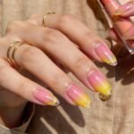 A hand with almond-shaped nails painted in a glossy pink-to-yellow ombré gradient. The tips are decorated with fine yellow glitter. The person wears gold rings and holds a peach-colored lip balm.