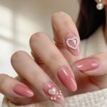 3d Nail Art Ideas That Look Luxe and Wearable Almond-shaped pink valentines nails with glossy polish, featuring pearl heart designs and delicate embellishments on the ring and index fingers.
