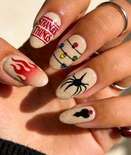 Beige almond-shaped nails with speckled detail, each featuring a unique Stranger Things design including red flames, the show's logo, multicolored Christmas lights, a black spider, and a shadowy shape with a red dot.