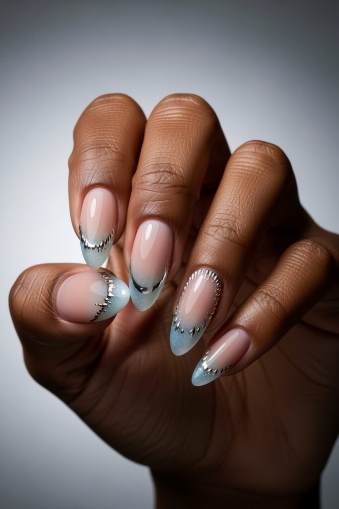 Hand with almond-shaped nails in a gradient icy blue ombré with metallic silver detailing along the tips, creating a sophisticated Winter Nails look.