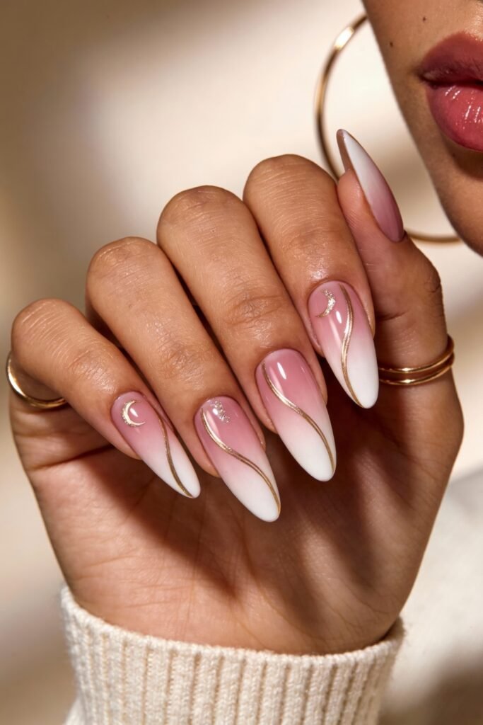 Pink and gold nails in a sharp almond shape, with a gradient pink to white base, gold swirls, and delicate crescent moon and star embellishments.