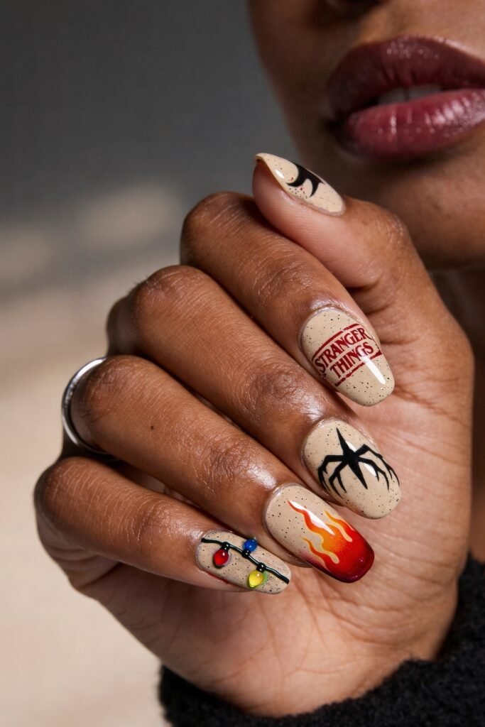 A close-up of a hand with speckled beige Stranger Things nails featuring the red logo, a black spider, Christmas lights, and a dramatic red-to-orange flame on the ring finger.