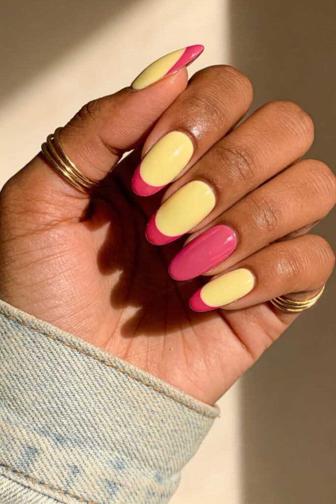 Pink and Yellow Nails That Feel Bright and Wearable 18 Almond nails in a glossy pink and yellow mix, featuring alternating color-blocking and curved tips. The person wears gold rings and a light denim jacket, and the nails are neatly polished with a bold contrast.