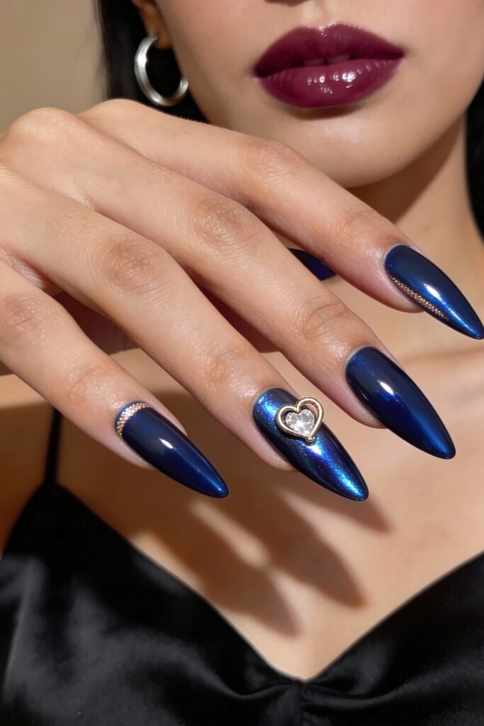 Long almond-shaped nails in a metallic midnight blue, featuring a gold-framed heart gem on the ring finger and rhinestone detailing for an elegant blue valentine nails finish.