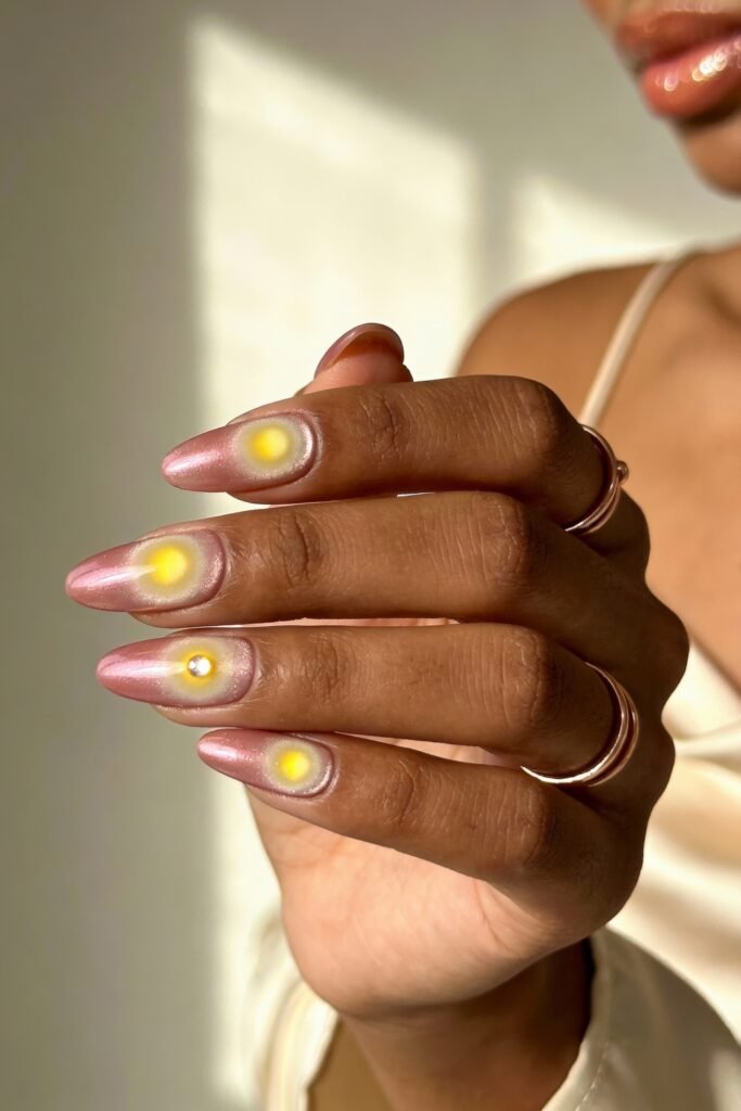 Pink and Yellow Nails That Feel Bright and Wearable 20 Almond-shaped nails painted in a shimmering soft pink, each featuring a glowing yellow dot in the center, giving a subtle cosmic or halo effect. One nail includes a small rhinestone embellishment. The hand is adorned with rose gold rings and a satin strap top is visible.