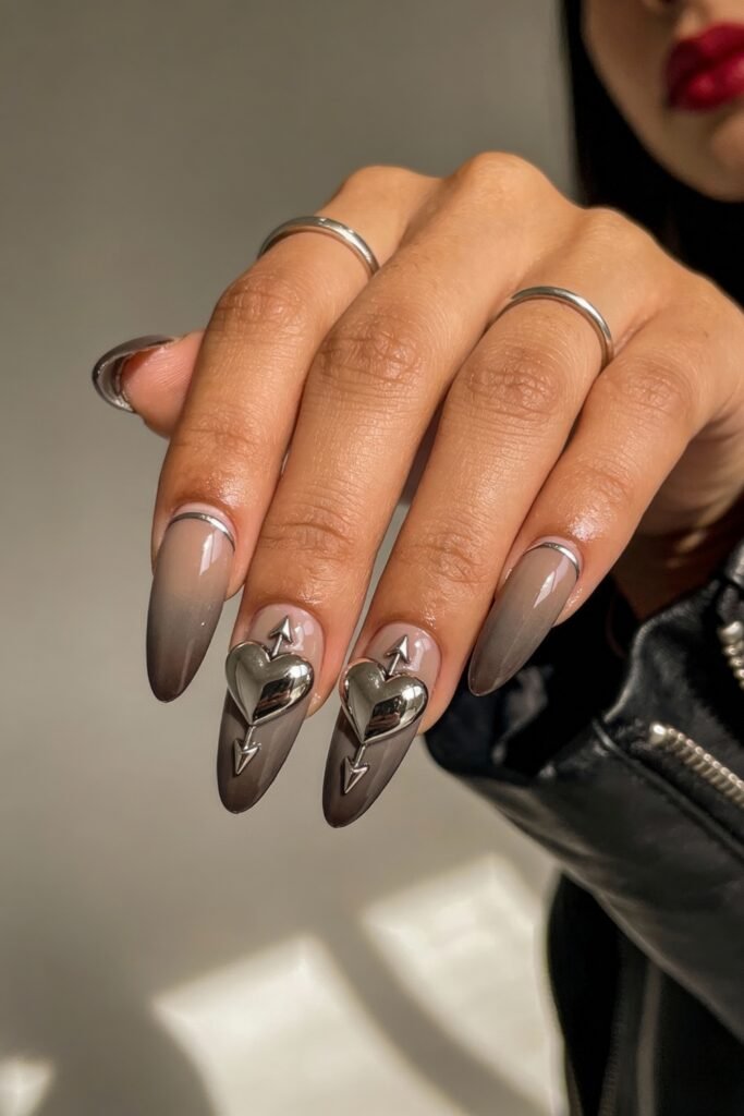 Gothic Valentines Nails That Feel Dark and Romantic 22 Almond-shaped gothic valentines nails in matte taupe ombré, featuring oversized metallic silver hearts pierced by vertical arrows for a bold romantic look.