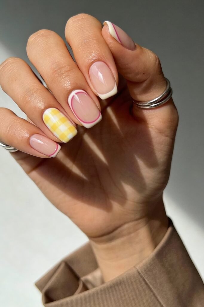 Pink and Yellow Nails That Feel Bright and Wearable 24 Short square nails with a nude base featuring white French tips outlined in pink, yellow gingham patterns, and minimalist line accents. The person wears silver rings and a tan blazer, with soft light casting shadows across the hand.