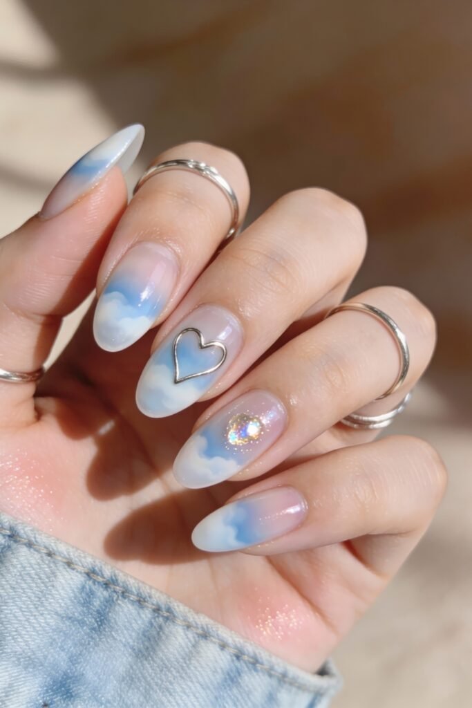 Soft almond-shaped nails with a blue sky and cloud motif, adorned with a metallic heart frame and a glittery gem accent, ideal for a whimsical blue valentine nails theme.