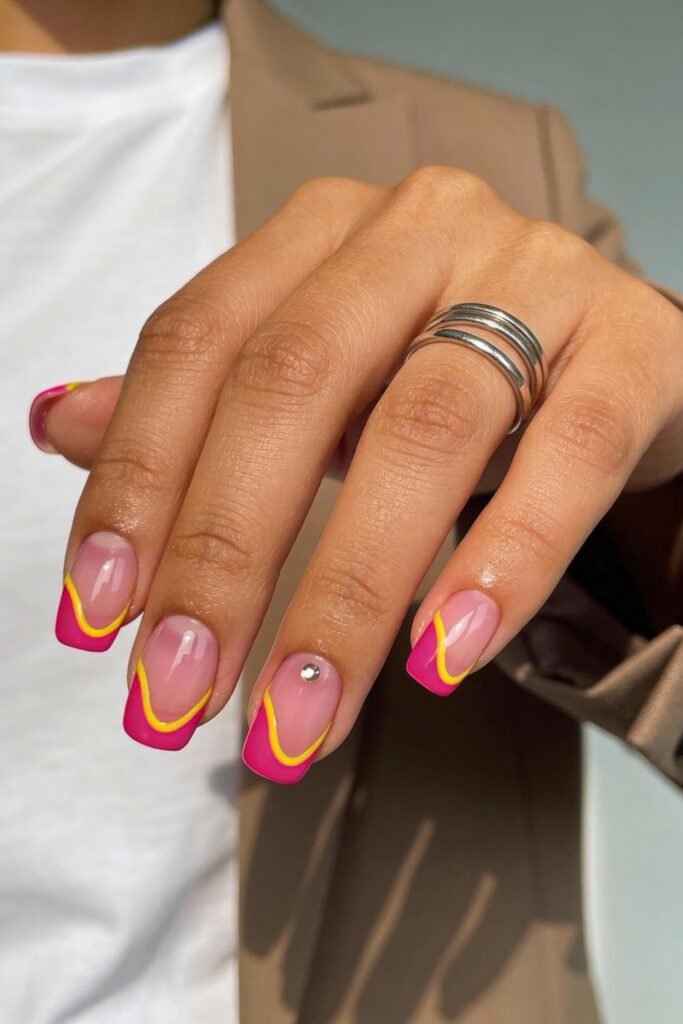 Pink and Yellow Nails That Feel Bright and Wearable 3 A hand with short square nails painted with a French tip design in hot pink and accented with wavy yellow lines. One nail features a small rhinestone. The person wears silver rings and a beige blazer.