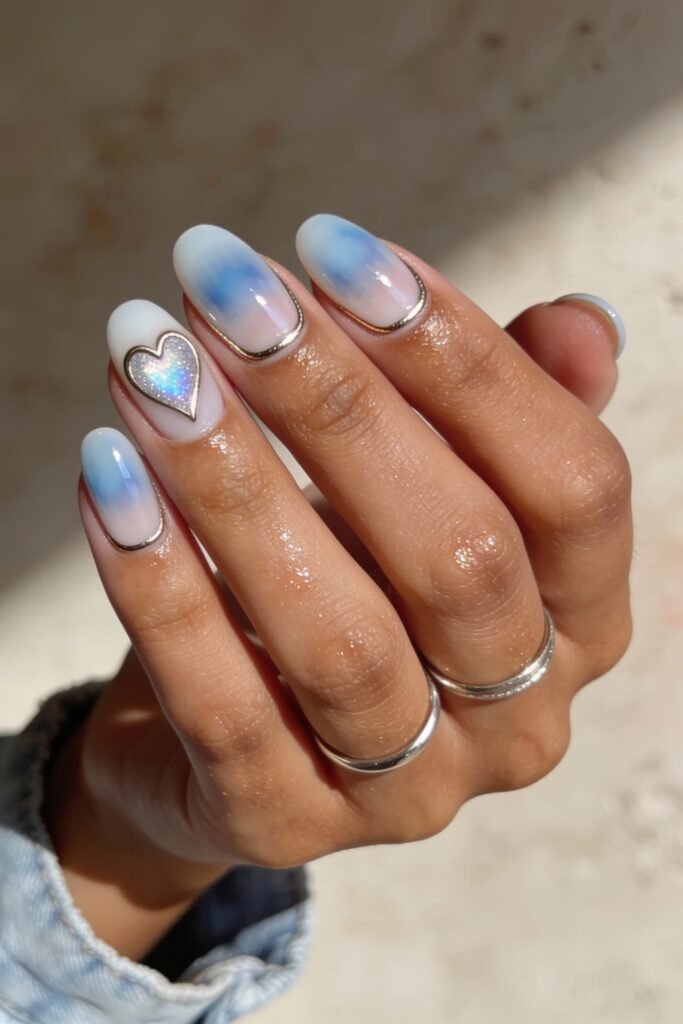 A set of rounded nails with a soft gradient of blue and white, framed in metallic chrome and highlighted with a holographic heart on the ring finger for a celestial blue valentine nails look.