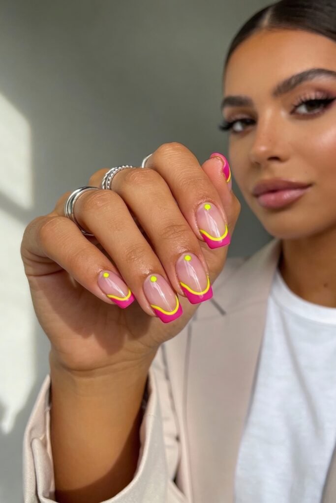 Pink and Yellow Nails That Feel Bright and Wearable 4 Bright pink French tip nails with yellow wavy accents and small neon dots at the center of the tips. The person wears silver rings and a white shirt under a beige blazer, with part of their face visible in the background.