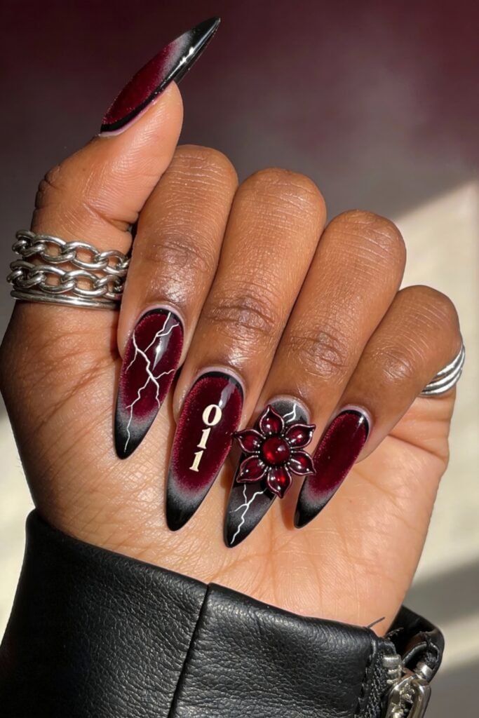 Burgundy and black ombré nails featuring lightning bolts, gold “011” lettering, and a sculpted Demogorgon flower with a red gem center, styled with stacked silver rings.