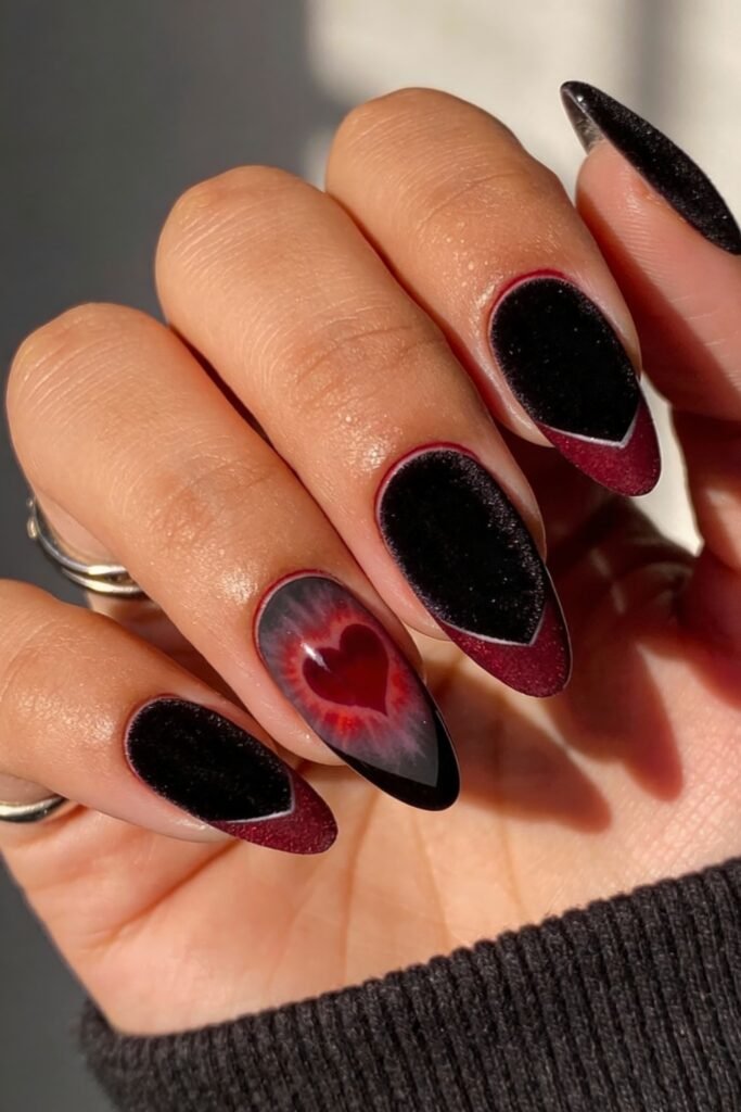 Gothic Valentines Nails That Feel Dark and Romantic 4 Almond-shaped gothic valentines nails with a velvet-textured black and burgundy base, accented by a glowing red heart on the ring finger.