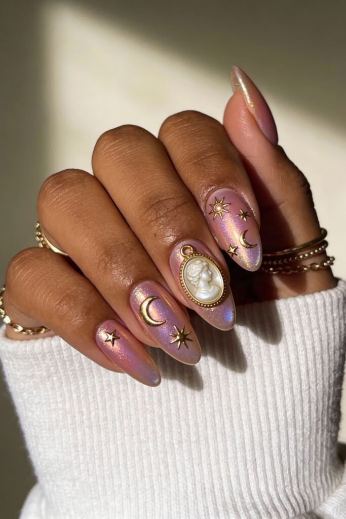 Iridescent pink and gold nails with celestial gold accents including crescent moons, stars, and a cameo-style face charm on the ring finger.