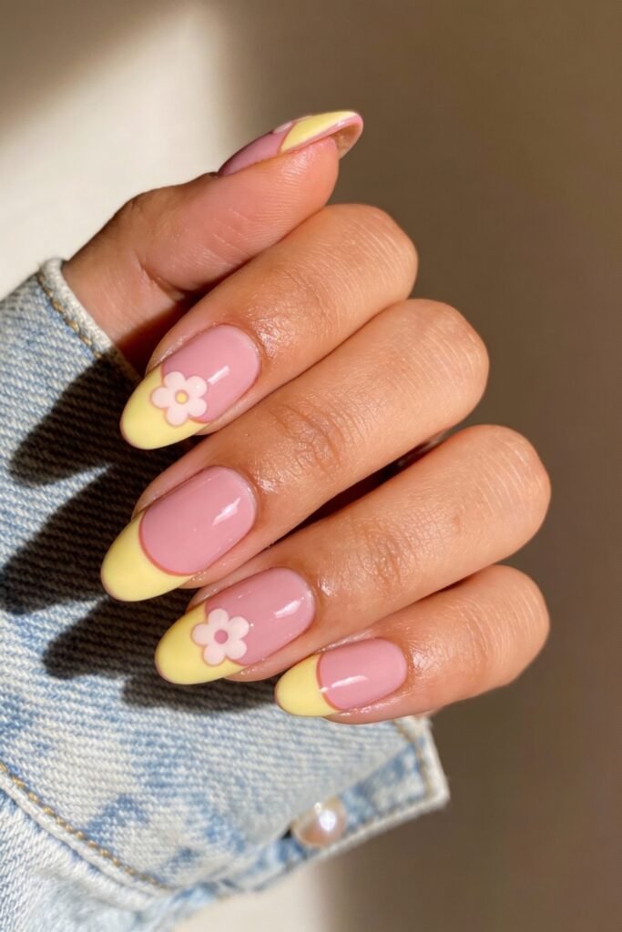 Pink and Yellow Nails That Feel Bright and Wearable 8 Oval-shaped nails with glossy pink polish and pastel yellow French tips. Each nail is accented with a single flower embellishment. The hand rests against a soft background, and the person wears a denim jacket.