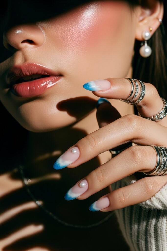 Hand with almond-shaped nails featuring blue and white ombré tips and small pearl embellishments, styled with rings and a soft winter glow.