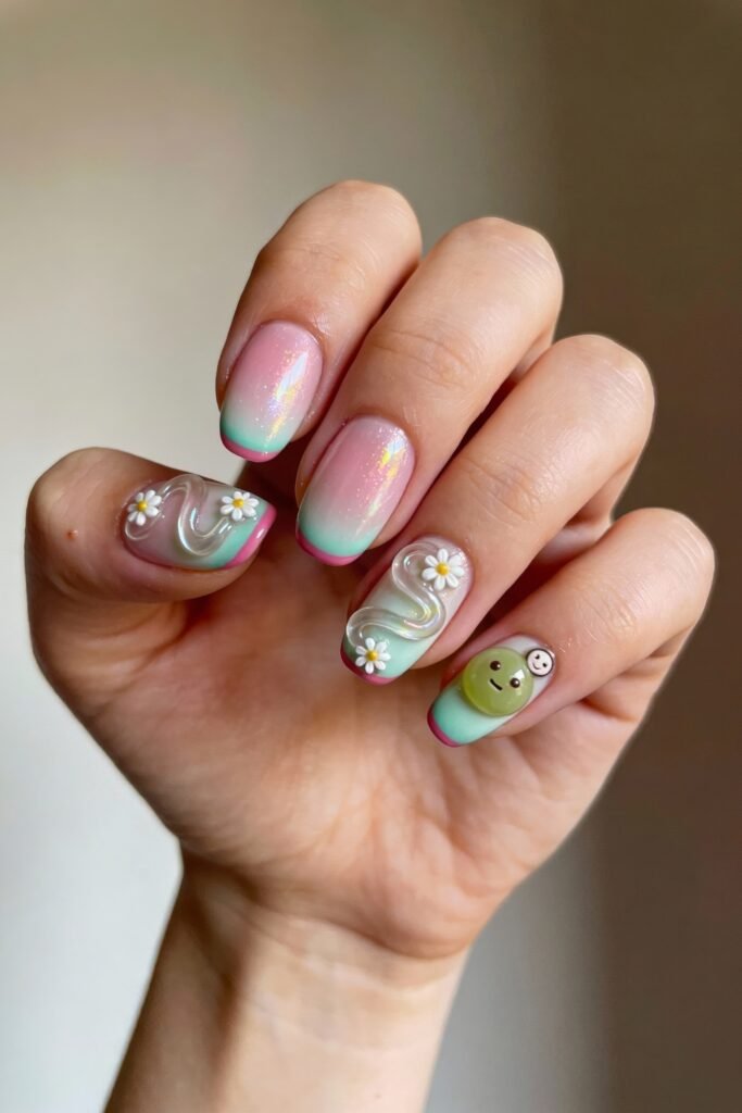 Short rounded nails with a soft pink and green gradient, enhanced with shimmer, 3D daisies, wavy accents, and a smiling green character on the pinky, creating a cheerful pink and green nails theme.