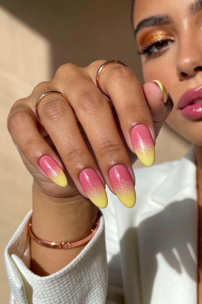 Pink and Yellow Nails That Feel Bright and Wearable 10 Almond-shaped nails with a glossy pink-to-yellow gradient design, enhanced with subtle glitter. The person wears rose gold rings and a bracelet, and their makeup features shimmering gold eyeshadow and glossy pink lips.