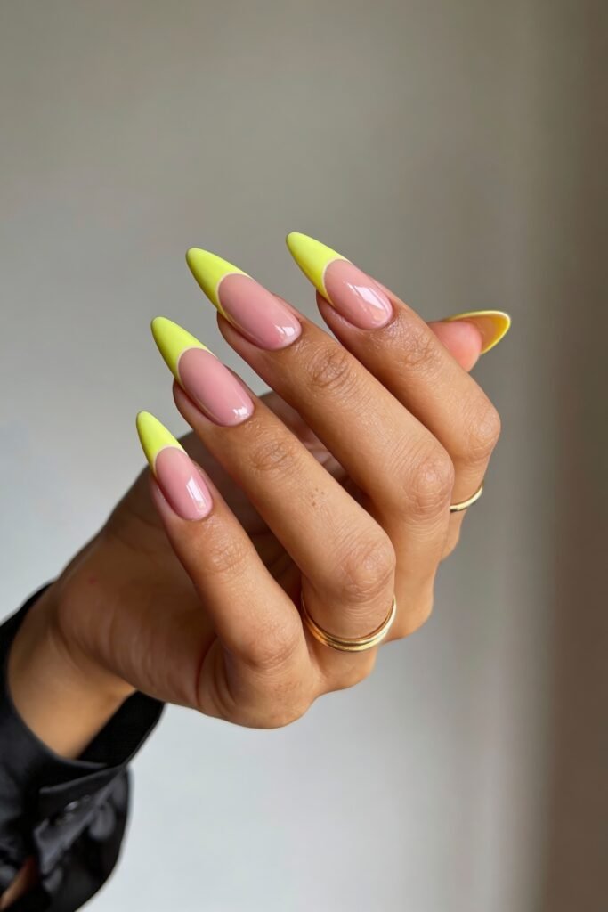 Pink and Yellow Nails That Feel Bright and Wearable 11 A hand with long almond nails painted in a minimalist reverse French tip style. The nails have a nude-pink base with bright neon yellow crescent-shaped tips. The person wears gold rings and a black outfit.