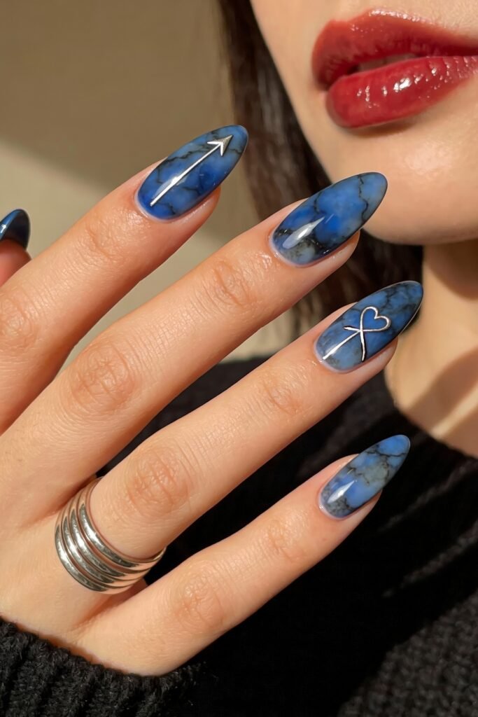 Almond-shaped nails with a blue marble effect, featuring silver metallic designs of a heart with an arrow and a directional arrow, blending edgy romance into a unique blue valentine nails style.