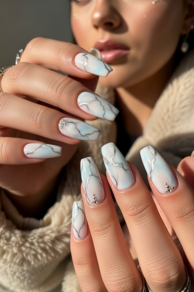 Hands with long square nails in a soft marble effect of white, grey, and icy blue, accented with tiny rhinestones, evoking a cool winter stone aesthetic.