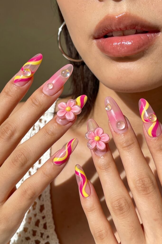 Pink and Yellow Nails That Feel Bright and Wearable 12 Playful, almond-shaped nails with swirling pink and yellow lines, clear bubble-like accents, and large 3D pink flower embellishments. The nail design is glossy and whimsical, and the person's lips and silver hoop earrings are visible in the background.