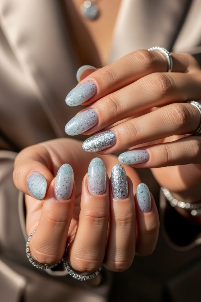 Hands with almond-shaped nails in a sheer icy blue polish topped with delicate silver glitter, including a couple of fully glittered accent nails, giving a sparkling winter effect.