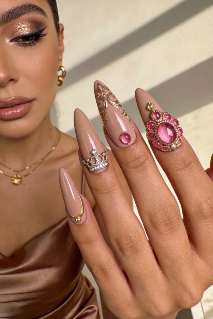 Long stiletto-shaped nude nails with regal pink and gold details, including rhinestone crowns and ornate metallic embellishments.