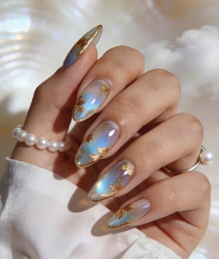 Goddess Nails iridescent blue chrome almond nails with gold leaf foil and glitter tips, styled with pearl jewelry in soft light