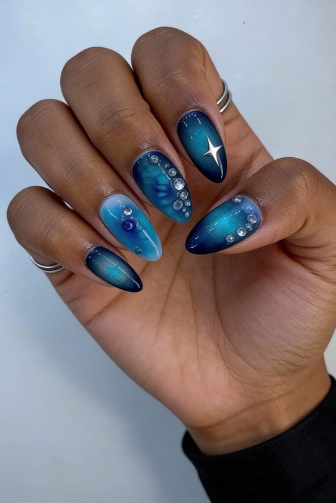Hand with blue gradient avatar nails blending teal and navy, adorned with rhinestones, galaxy-inspired shimmer, and a chrome star accent, creating a celestial and futuristic vibe.