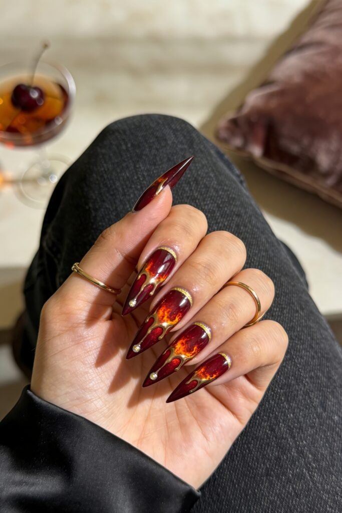 Fire Nails That Look Expensive and Feel Effortless 1 Burgundy stiletto nails with fiery orange marbling and metallic gold drip details, hand with gold rings