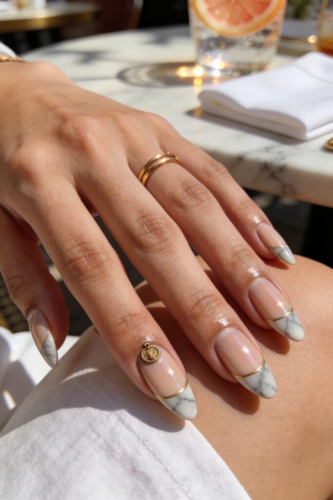 Medium almond Goddess Nails displaying a sheer base with diagonal white marble tips divided by a thin gold line and a small gold medallion charm near the cuticle