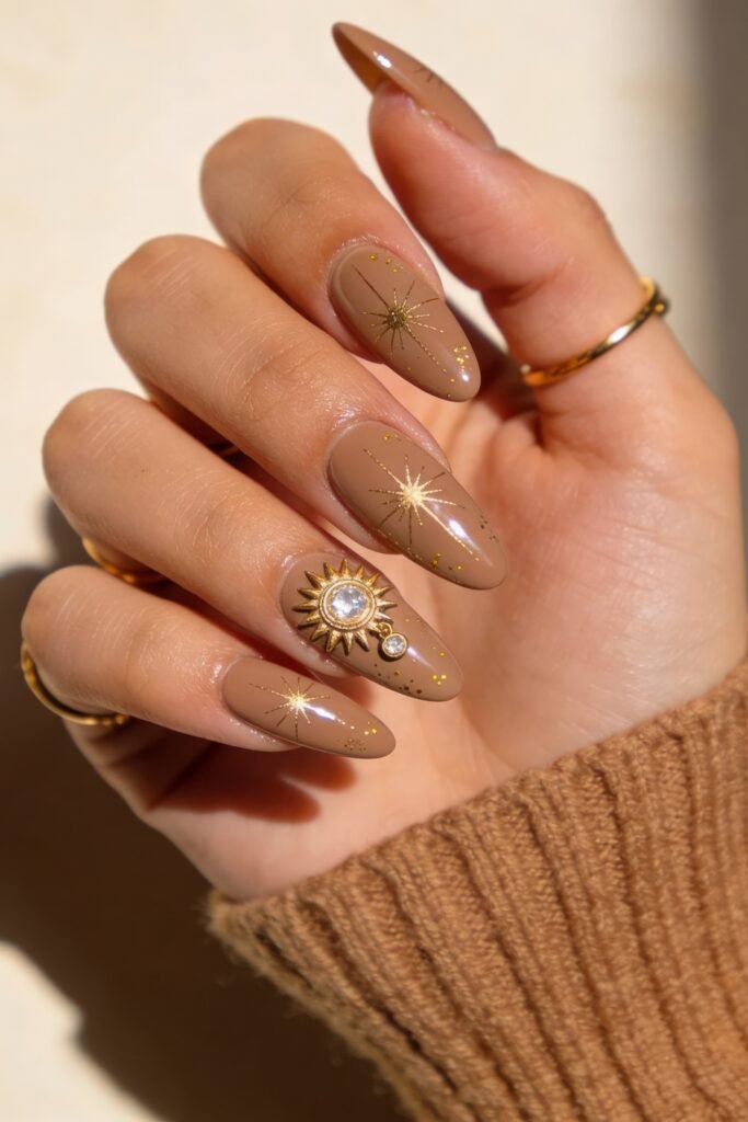 Nude mocha almond nails with gold starburst line art, scattered gold flecks, and a sun charm with rhinestones