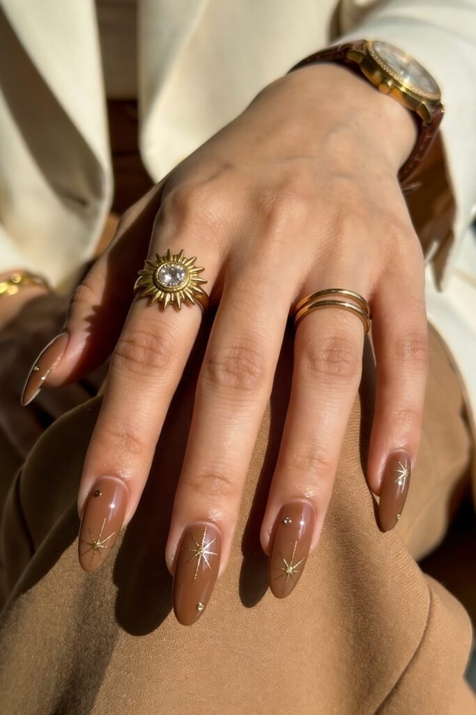 Hand resting on tan fabric showing glossy taupe nails with gold starburst accents and tiny studs, styled with gold rings and watch