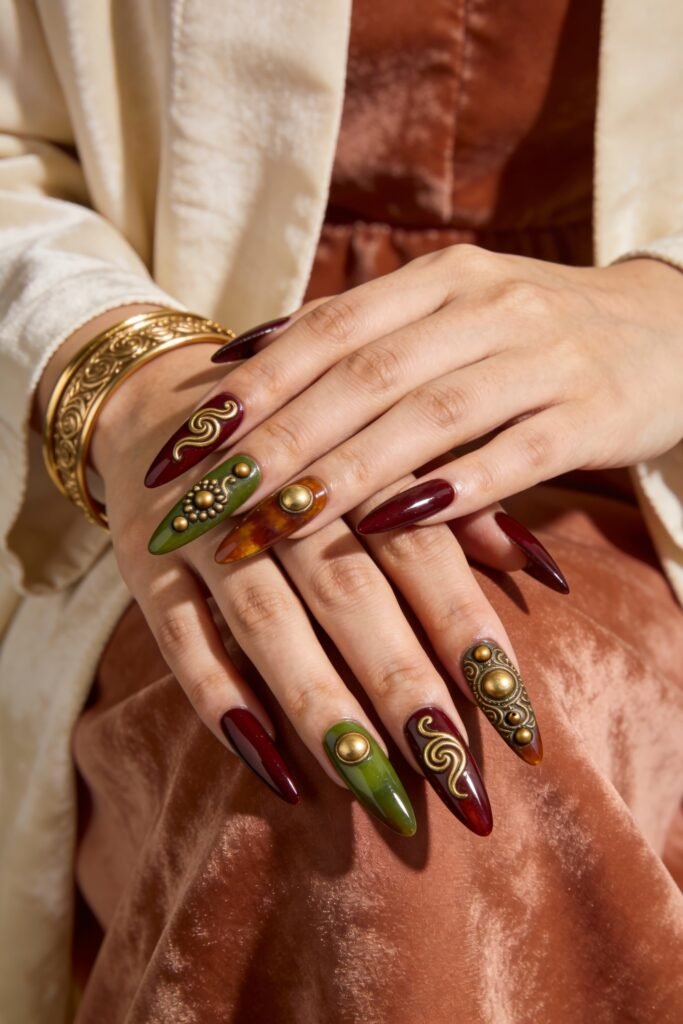 Two hands with long stiletto nails in burgundy, olive green, and amber tortoise, featuring raised gold embellishments and studs