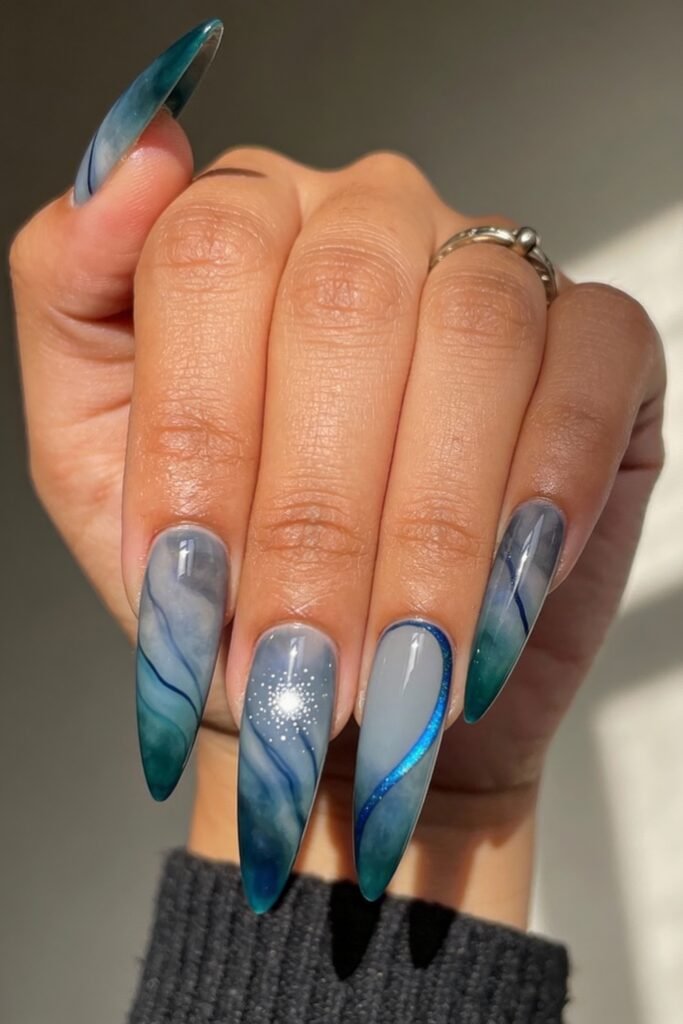 Glossy, pointed avatar nails in blue and gray tones, adorned with swirling ocean-like patterns, a glowing starburst accent, and a metallic blue wave line for a dreamy aquatic design.