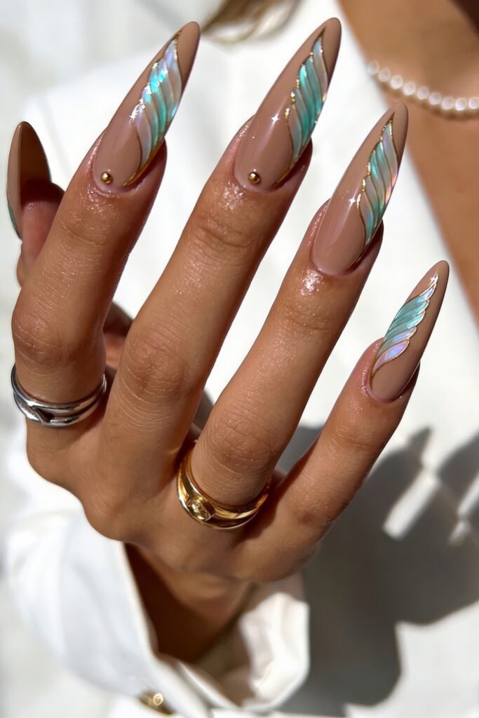 Nude stiletto nails with iridescent teal wing tips, fine gold outlines, and tiny gold studs in bright sunlight