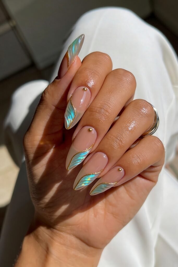 Nude almond nails with shimmering aqua leaf-tip design, gold trim, and small gold studs, hand posed on white fabric