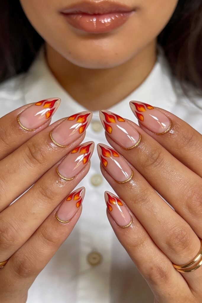 Fire Nails That Look Expensive and Feel Effortless 12 Nude stiletto Fire Nails with red-and-orange flame art and a delicate gold outline near the cuticles