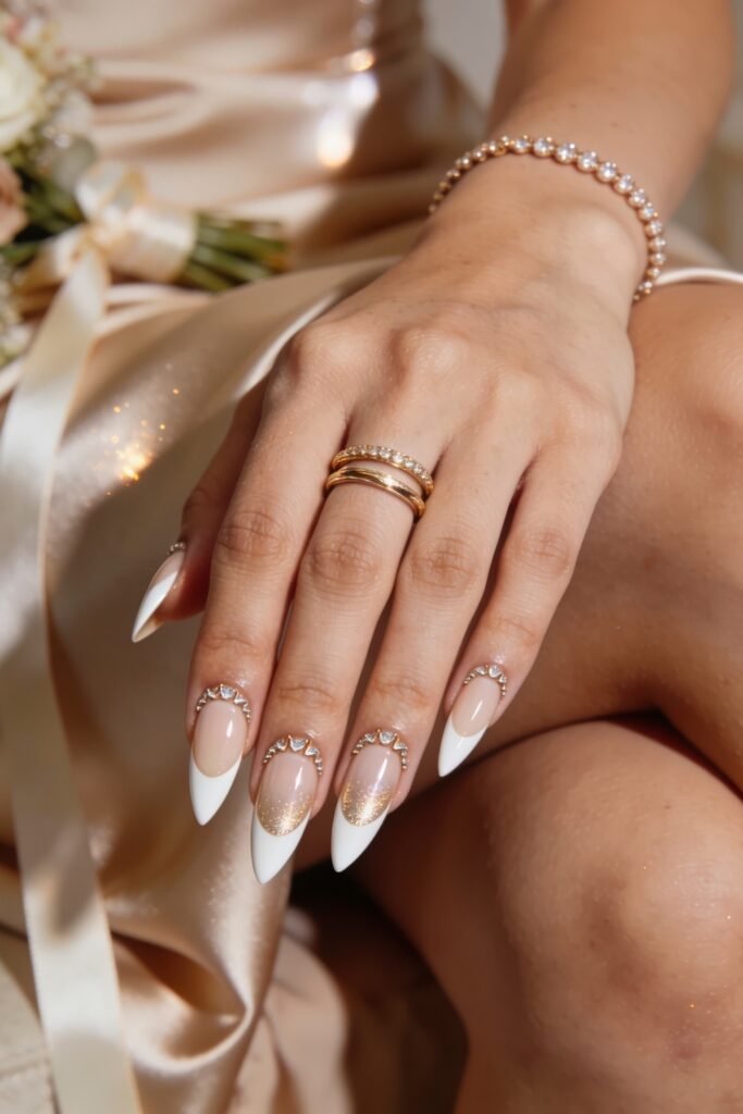 Goddess Nails bridal white French tips with rhinestone cuticle accents and gold shimmer detail, hand resting on satin fabric