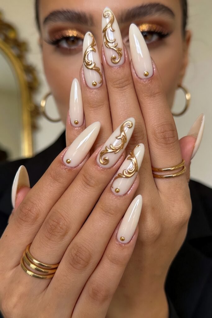 Cream ombré almond nails with ornate gold scrollwork and small gold studs, hands posed in front of glam makeup look