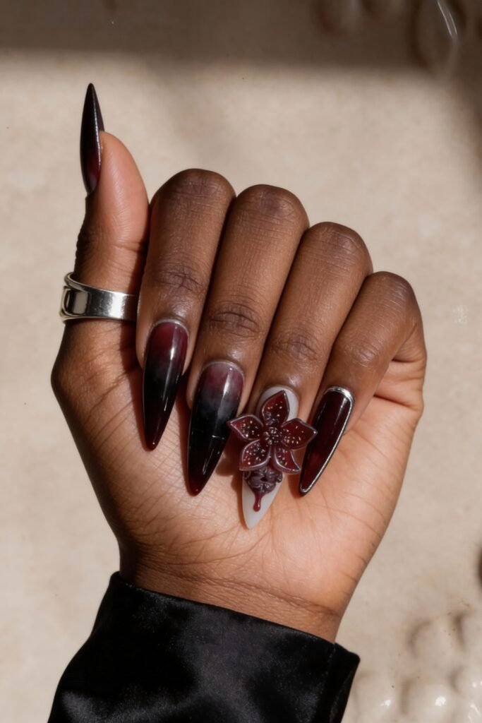 Stiletto nails with a Stranger Things theme, combining a deep burgundy fade, glossy black, and a sculpted Demogorgon flower dripping red onto a pale gray accent nail.