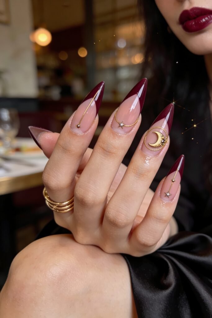 A hand modeling long pointed Goddess Nails featuring a nude base with dark burgundy V-tips and minimalist gold constellation lines and moon charms