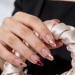 Close-up of a hand with long almond nude pink nails decorated with pink, burgundy, and gold glitter confetti, holding a cream satin pouch