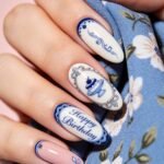 Close-up of a hand with long porcelain-inspired blue and white nails, featuring delicate floral scrollwork, a cake illustration, and Happy Birthday lettering against blue floral fabric