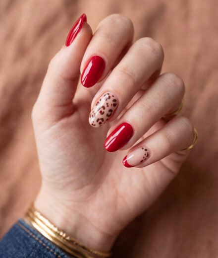 Hand with glossy red almond nails featuring leopard print accent nails and a red French tip against a warm blurred background